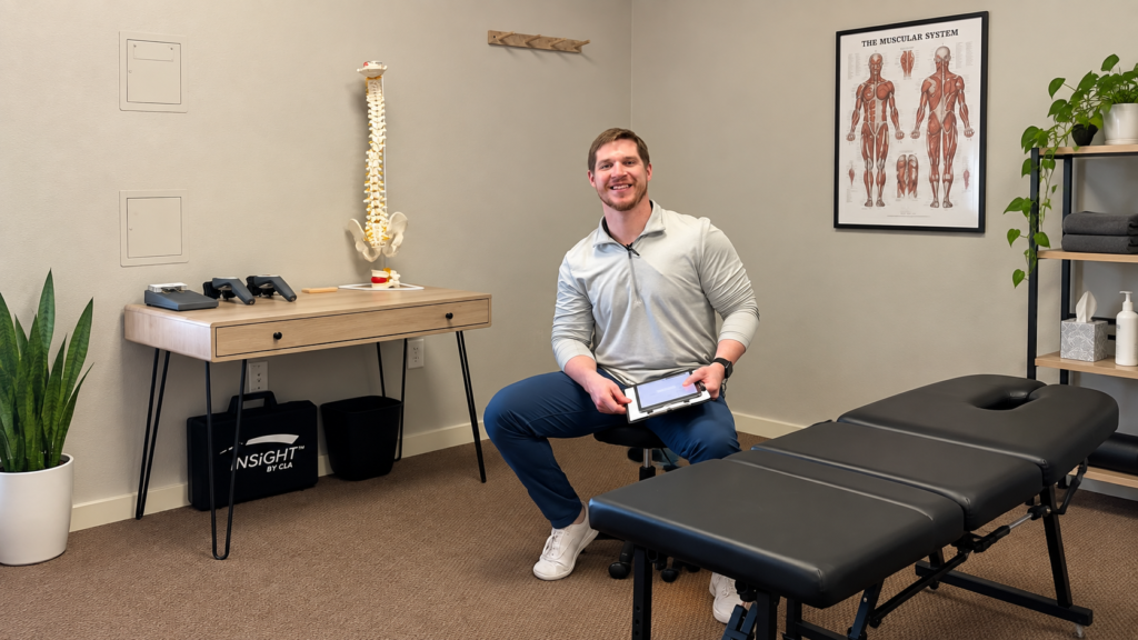 Dr. Cameron Williams in a treatment room at Radiant Chiropractic with a tablet, spine model, and CLA INSiGHT scanning equipment visible nearby