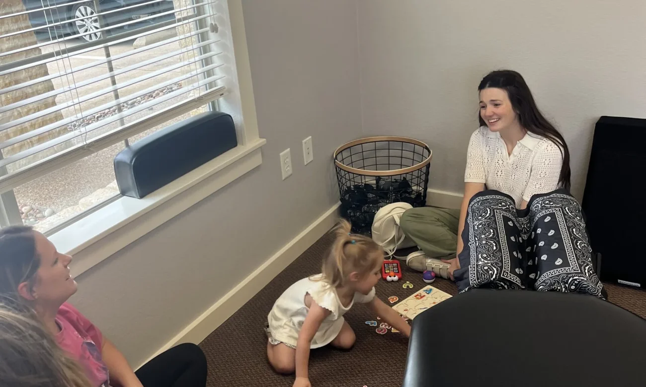 A toddler plays with a puzzle on the floor of a chiropractic room while women sit nearby and watch.