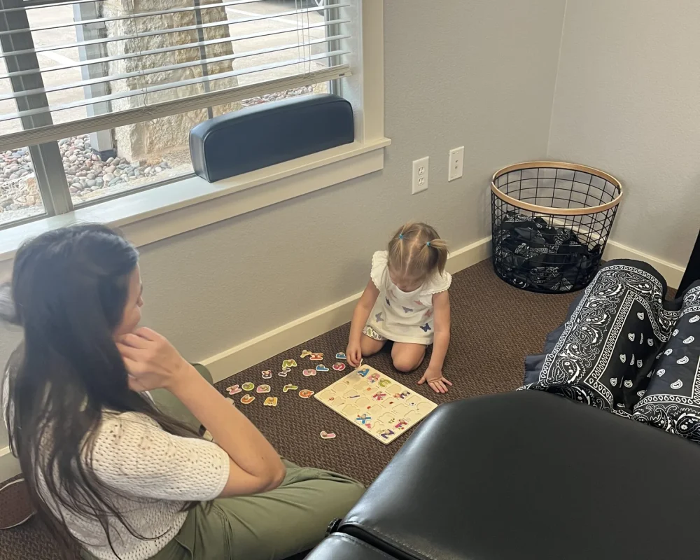 A toddler plays with an alphabet puzzle on the floor of a chiropractic treatment room next to a woman and adjusting table.