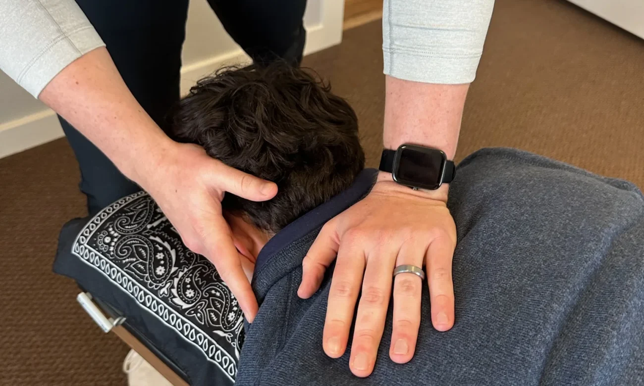 Chiropractor placing hands on a patient's neck and upper back while they rest face down on an adjusting table.