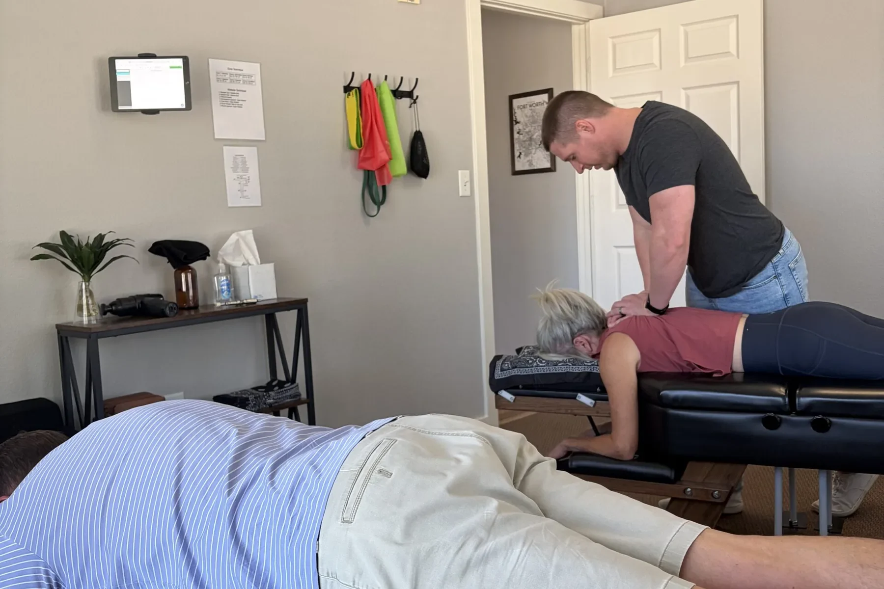 A chiropractor adjusts a woman's back on a treatment table while another patient rests on an adjacent table.