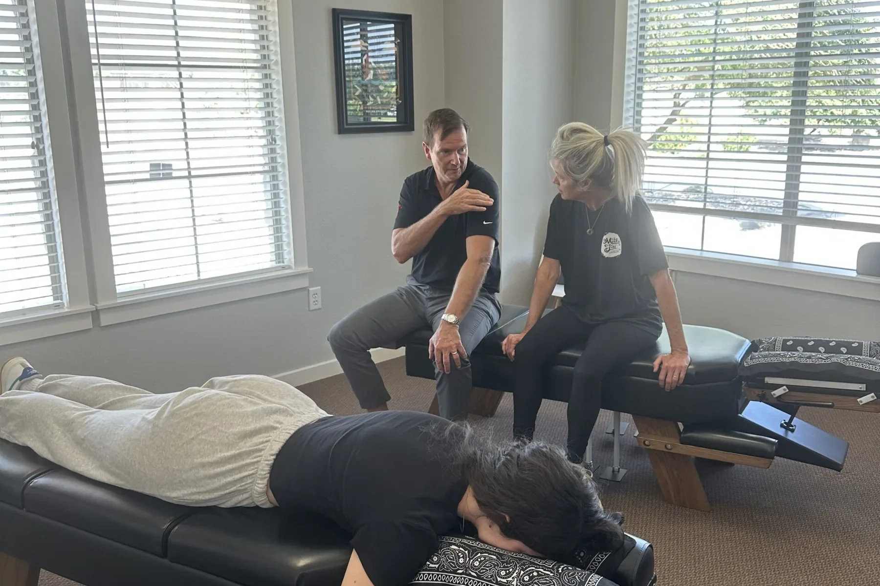 Chiropractor discussing care with a woman on an adjustment table while another person rests face down on a nearby table.