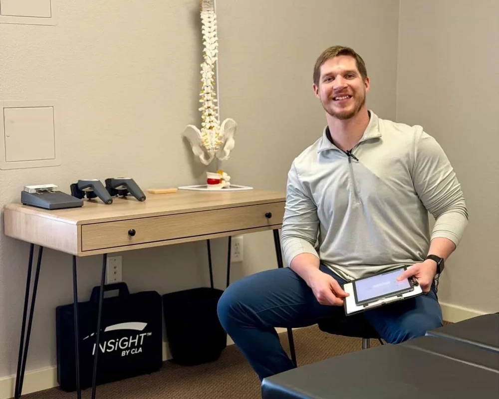 Dr. Cameron Williams seated in an examination room holding a tablet beside a desk with a spine model.