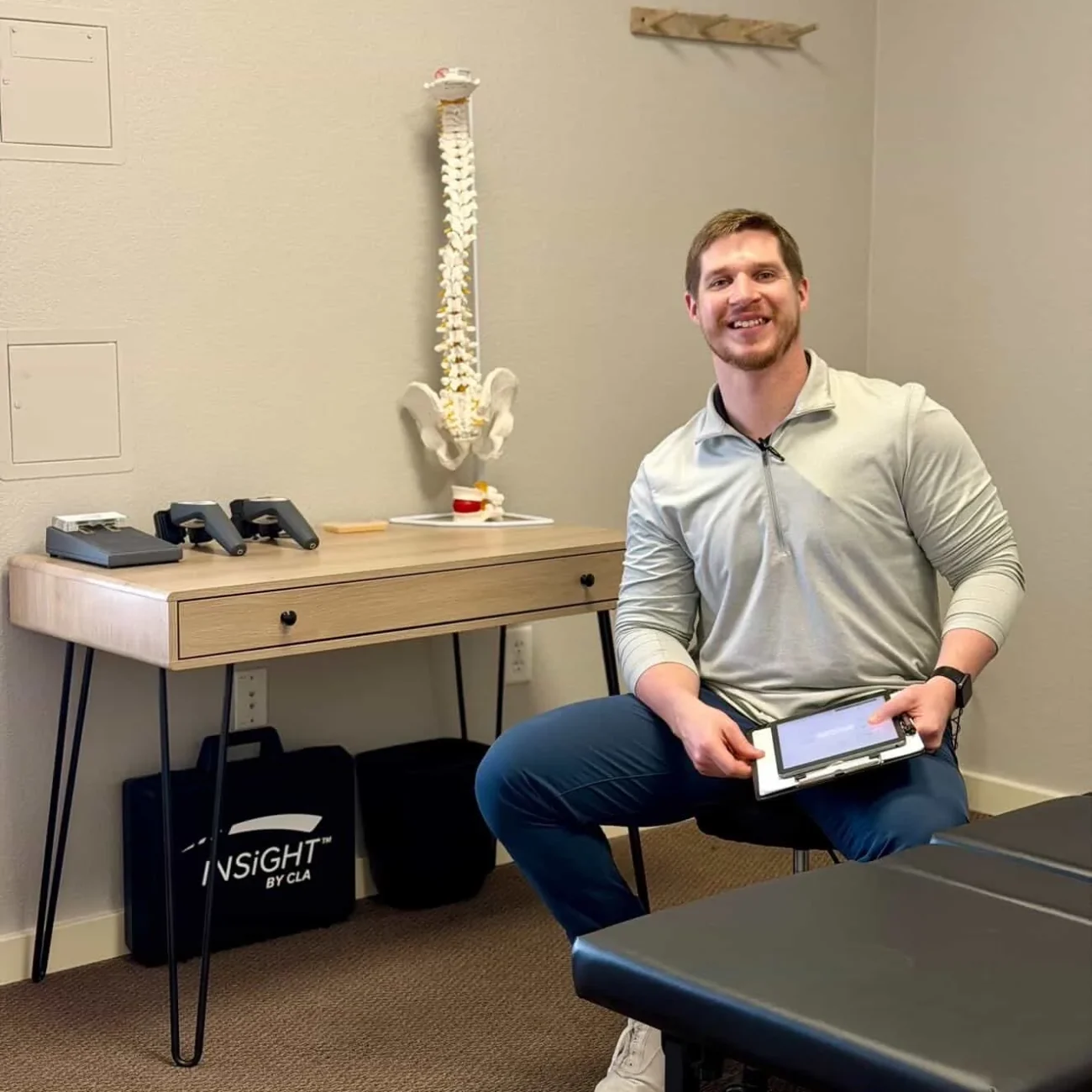 Dr. Cameron Williams seated in an examination room holding a tablet beside a desk with a spine model.