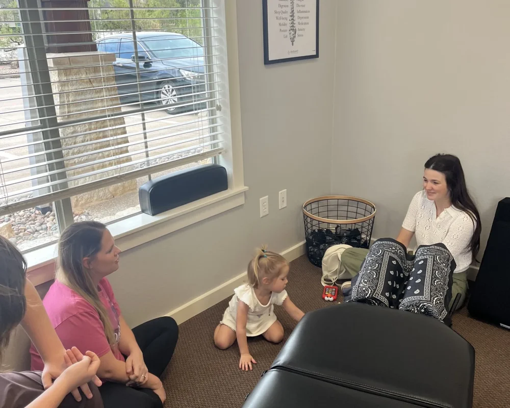 A chiropractor sitting on the floor with a mother and young child during an informal consultation in a treatment room.