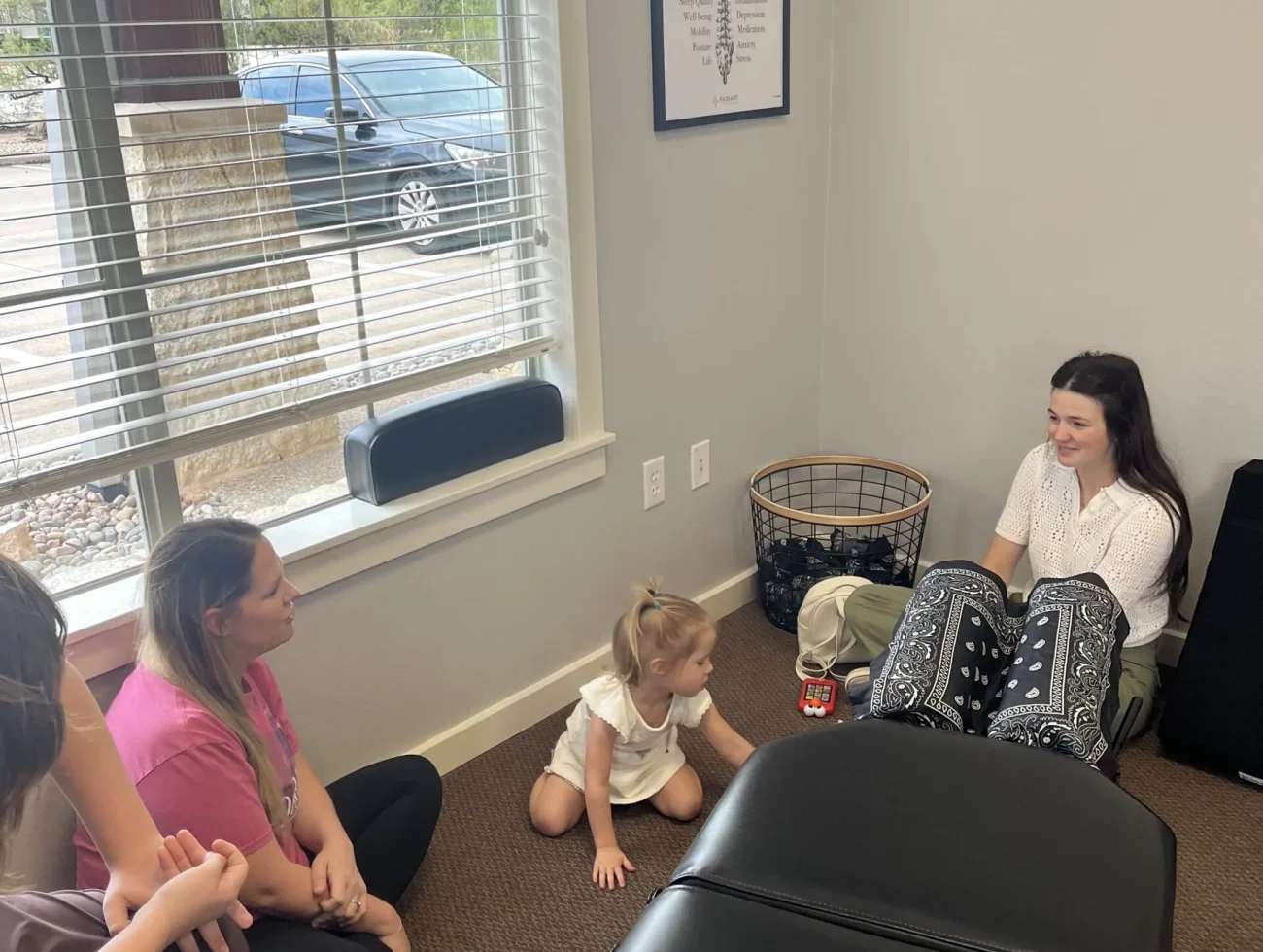 A chiropractor sitting on the floor with a mother and young child during an informal consultation in a treatment room.