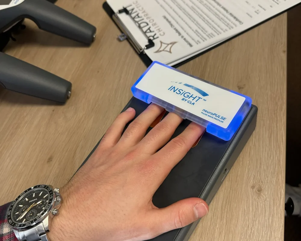 Patient's hand resting on a CLA INSiGHT neuroPULSE scanning device beside Radiant Chiropractic paperwork.