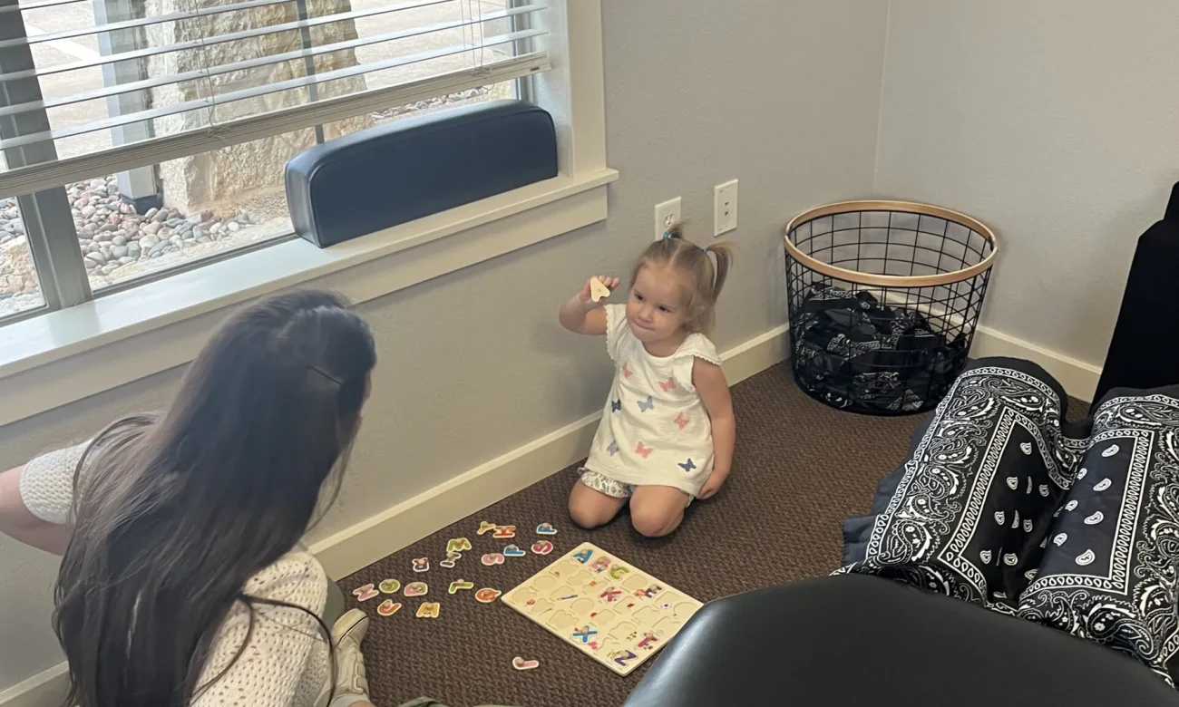 Toddler sitting on the floor playing with an alphabet puzzle next to a woman and an adjusting table in a chiropractic office.