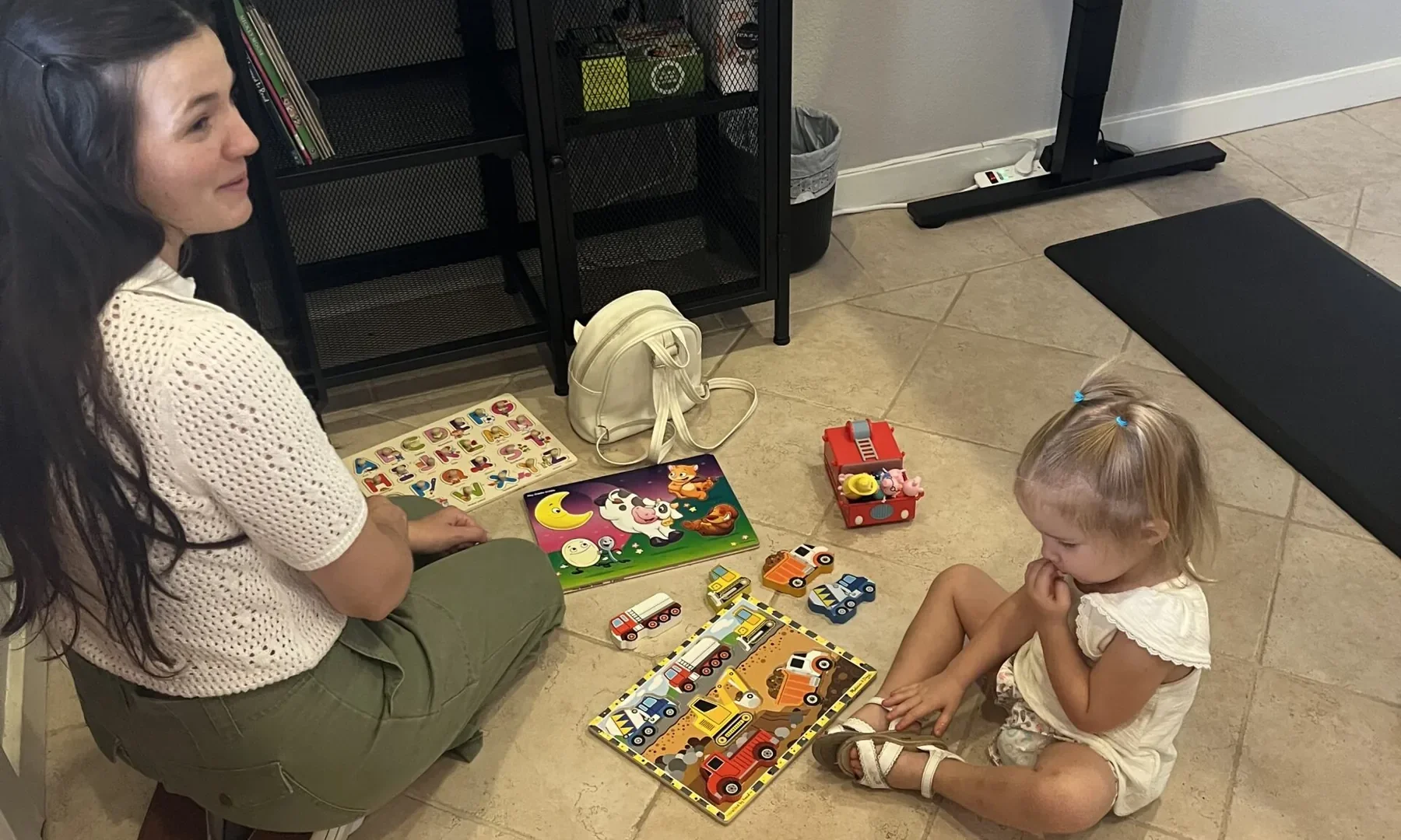 A woman and a young child sit on the floor playing with wooden puzzles and toy cars in an office waiting area.
