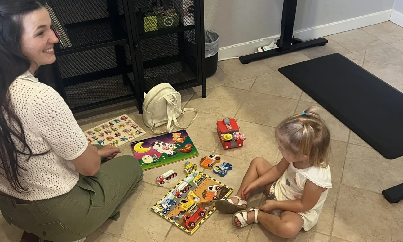 Woman and young child sitting on a tiled floor playing with wooden puzzles and toy trucks in an office setting.