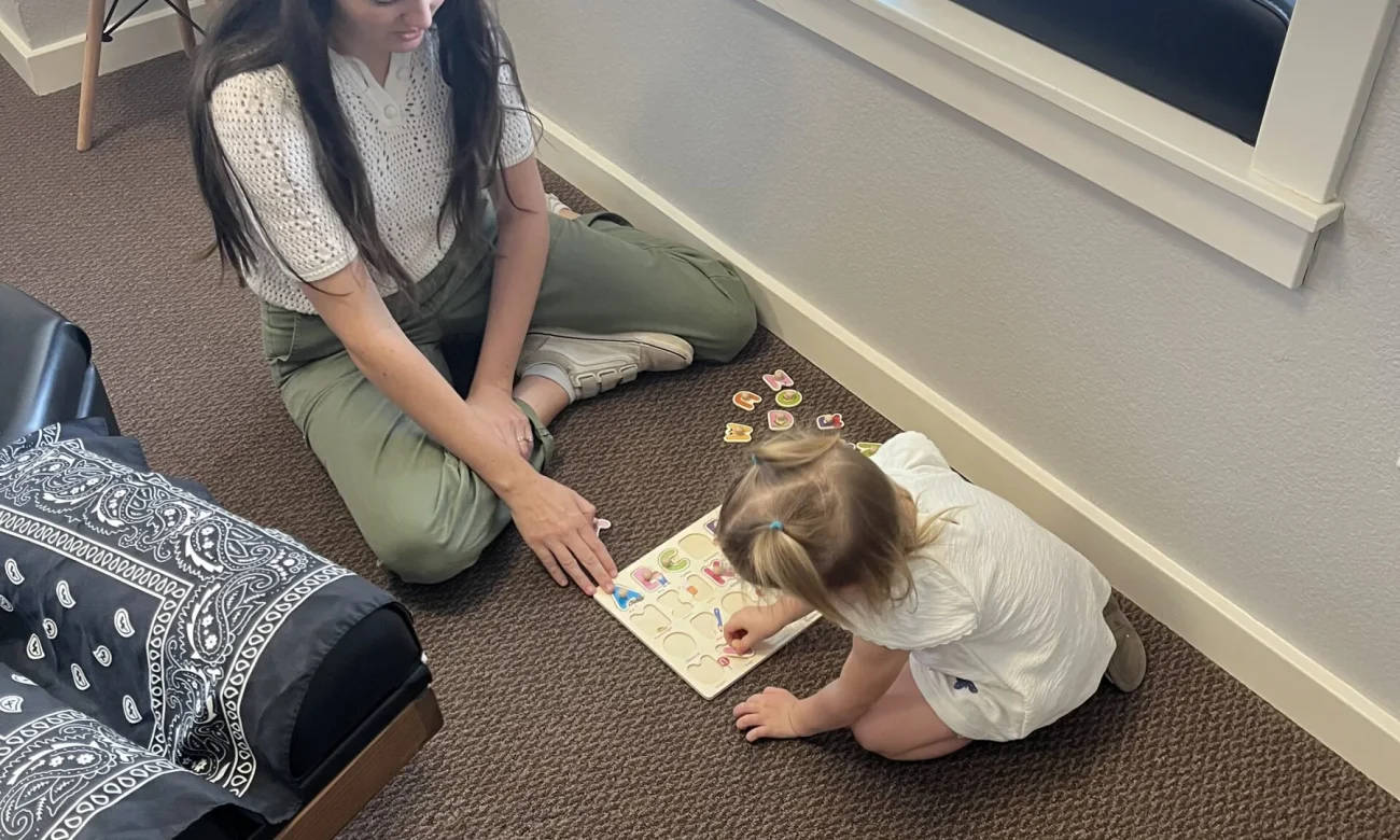 A woman and a toddler girl sit on the floor playing with a wooden alphabet puzzle inside a chiropractic office.
