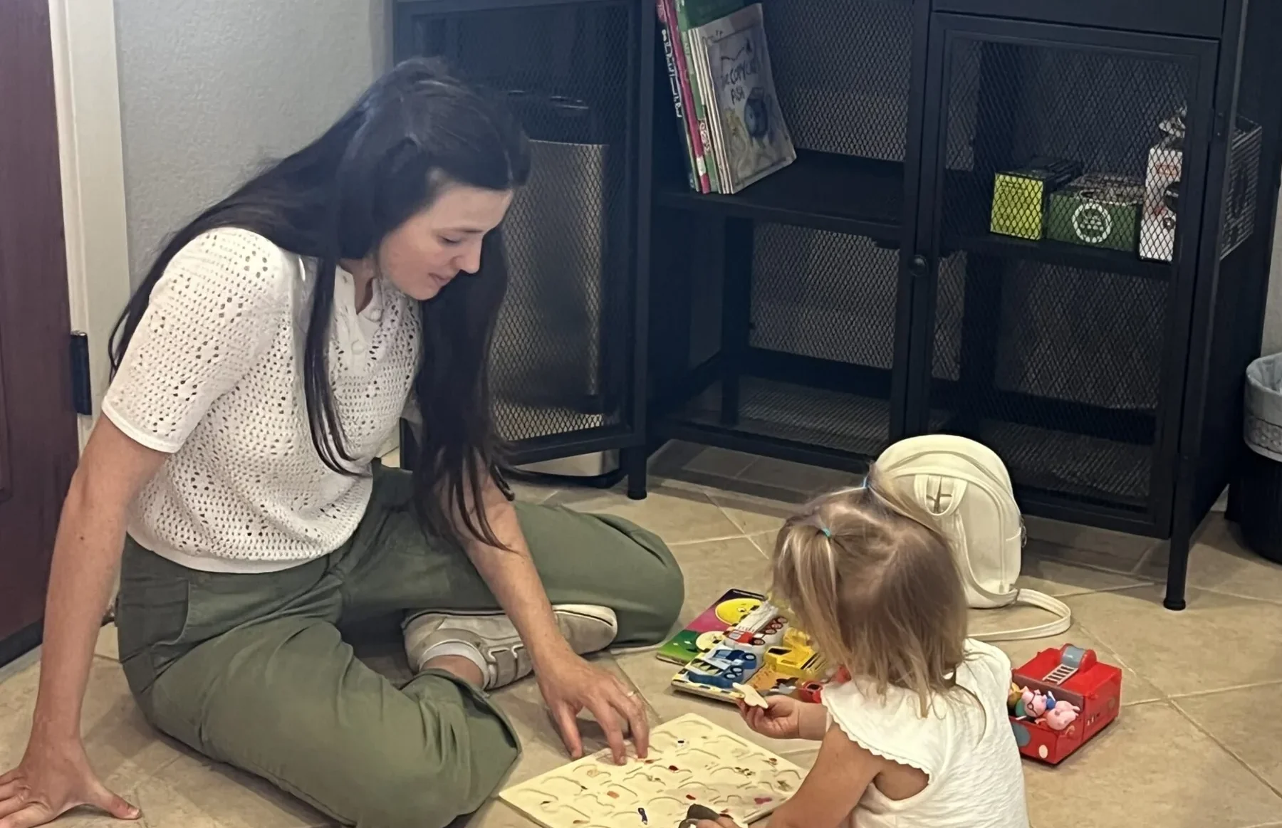 A woman sits on the tiled floor playing with an alphabet puzzle alongside a toddler in an office waiting area.